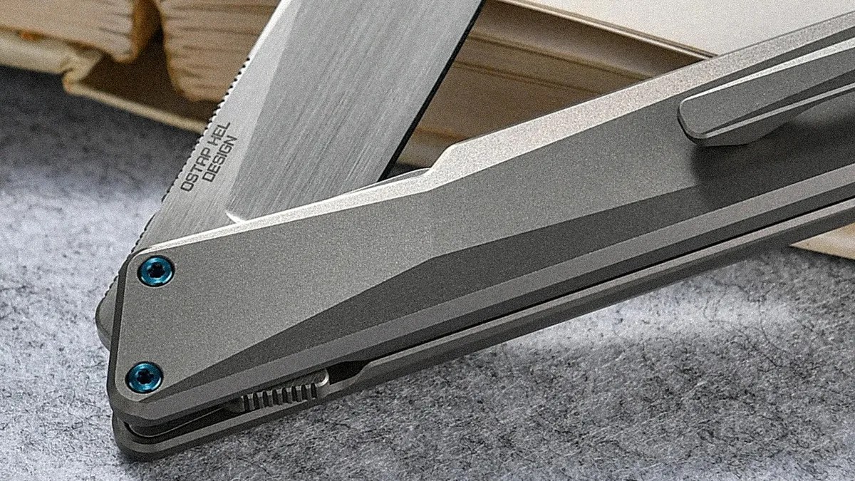 Close-up of a gray folding knife with blue screws and "OSTRO HEL DESIGN" engraved on the blade.