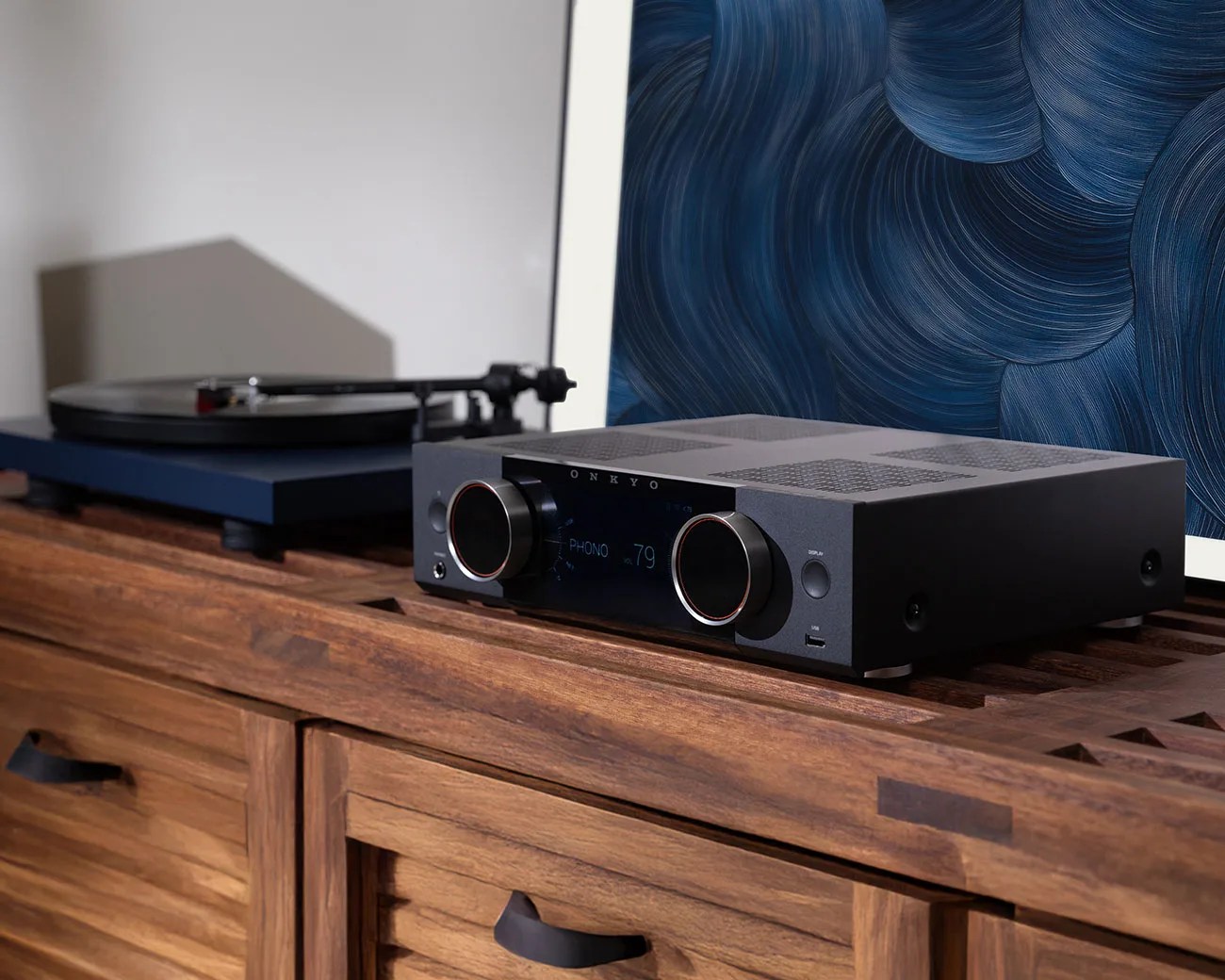 Black Onkyo stereo receiver with digital display and two large knobs on a wooden cabinet next to a blue turntable.