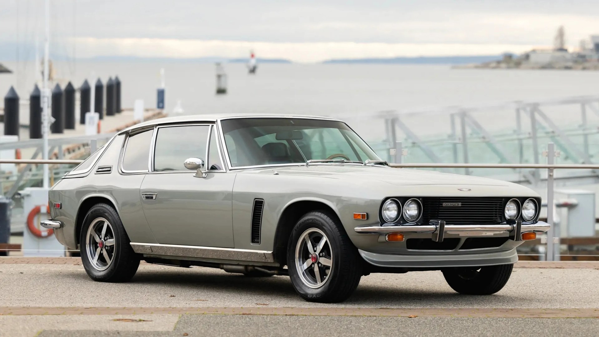 Silver classic Jensen Interceptor car parked near a waterfront with chrome trim and black alloy wheels.