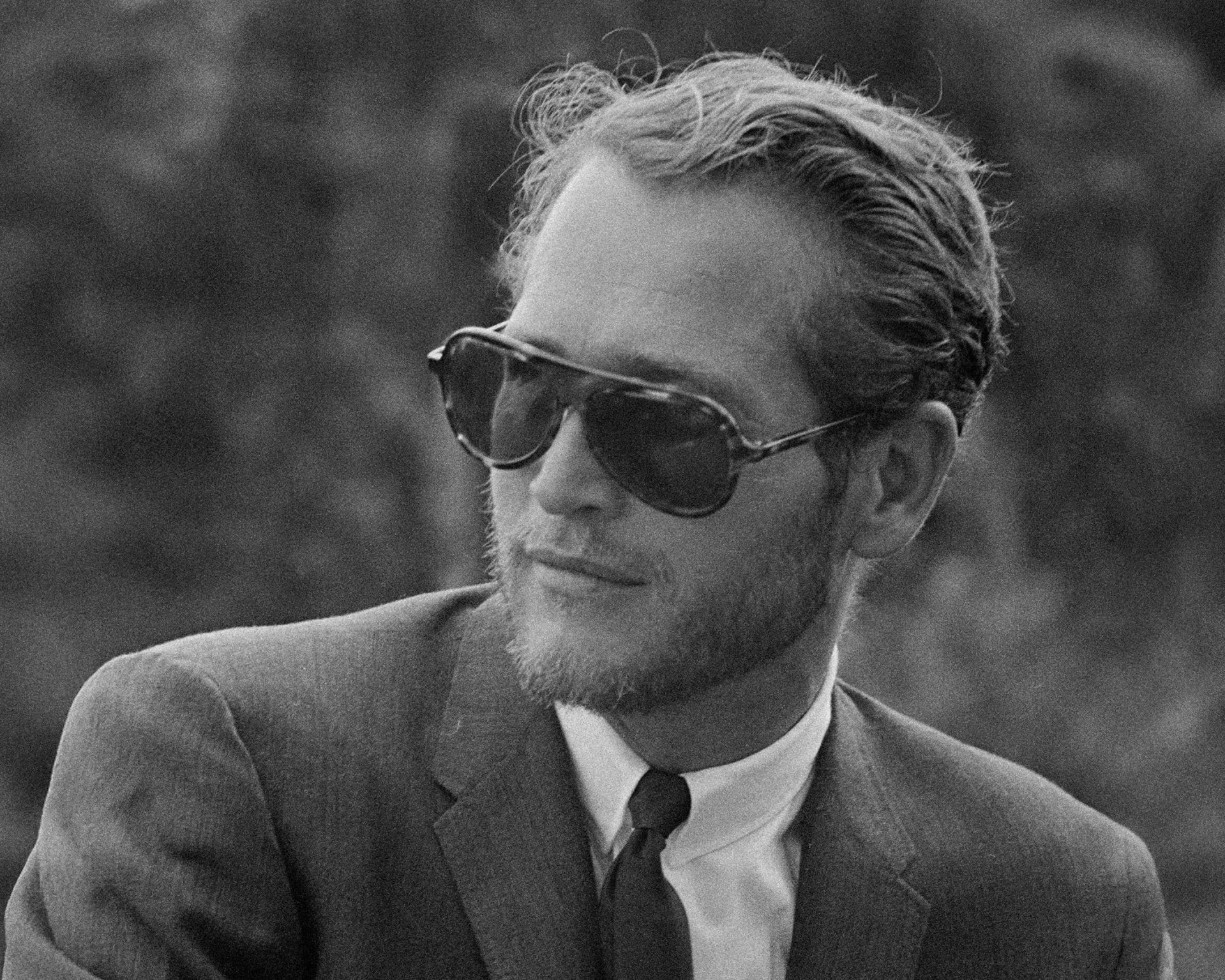 Man with short wavy hair and beard wearing dark aviator sunglasses, a suit jacket, white shirt, and tie.