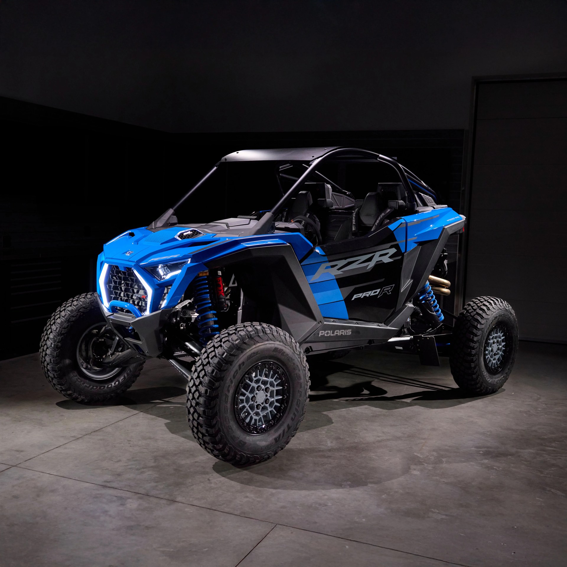 Blue and black Polaris RZR Pro R off-road vehicle with large knobby tires in a dimly lit garage.