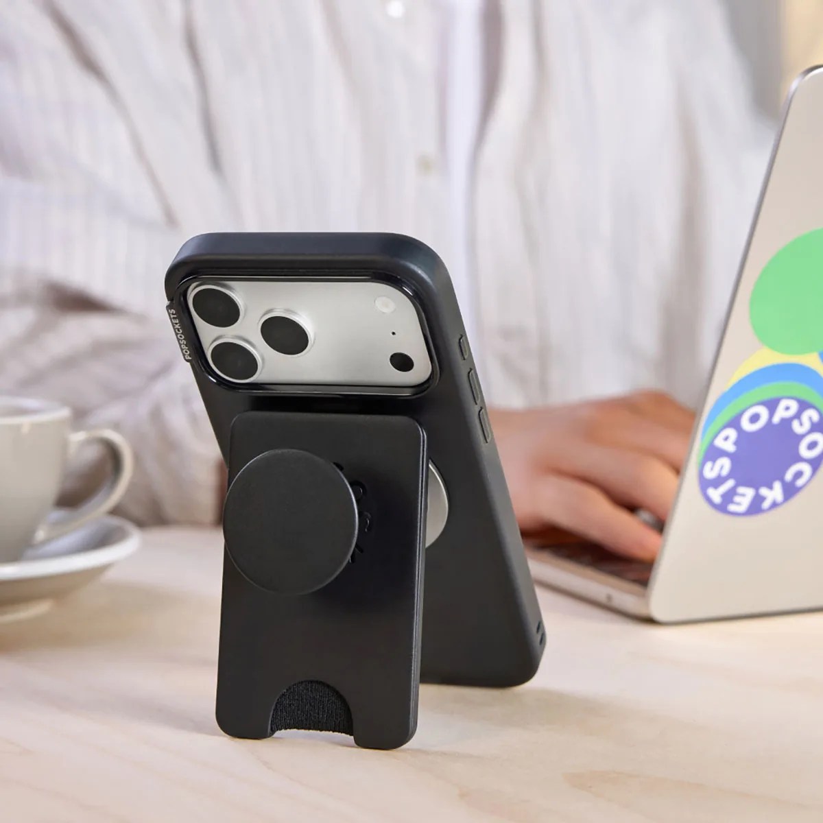 Black smartphone with a black PopSockets grip and stand attached, placed on a wooden surface near a laptop and a cup.