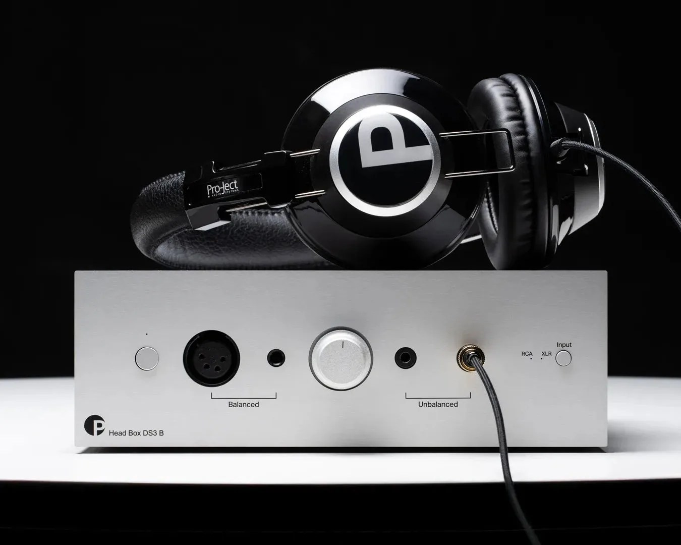 Black Pro-Ject Audio Systems headphones resting on a silver Head Box DS3 B headphone amplifier with input and volume controls.