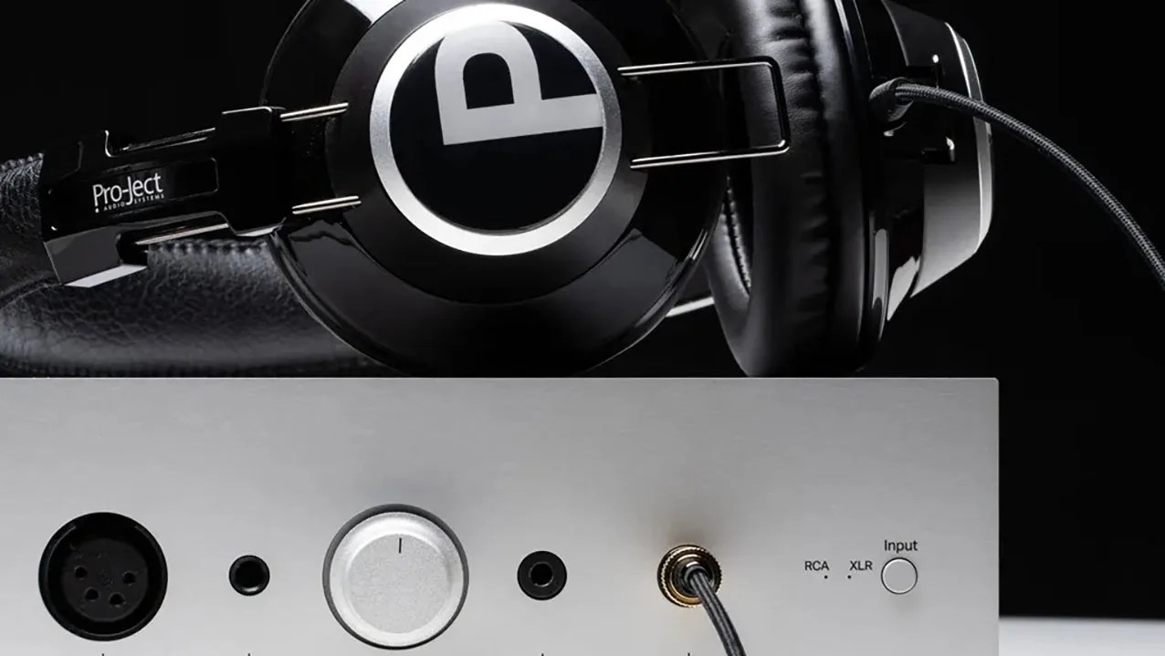 Black Pro-Ject Audio Systems headphones resting on a silver audio amplifier with input selector and cable connections.