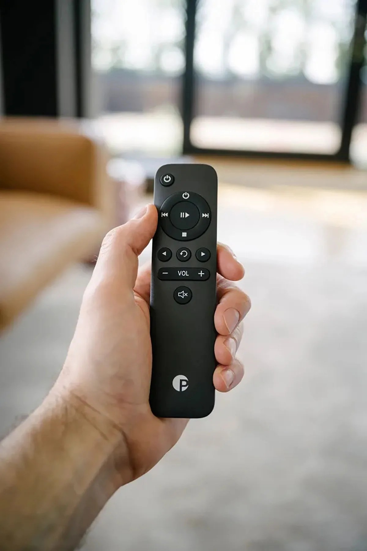 Hand holding a black remote control with playback and volume buttons in a living room.