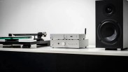 Silver audio amplifier and streaming device stacked on a white surface between a black turntable and a black speaker.