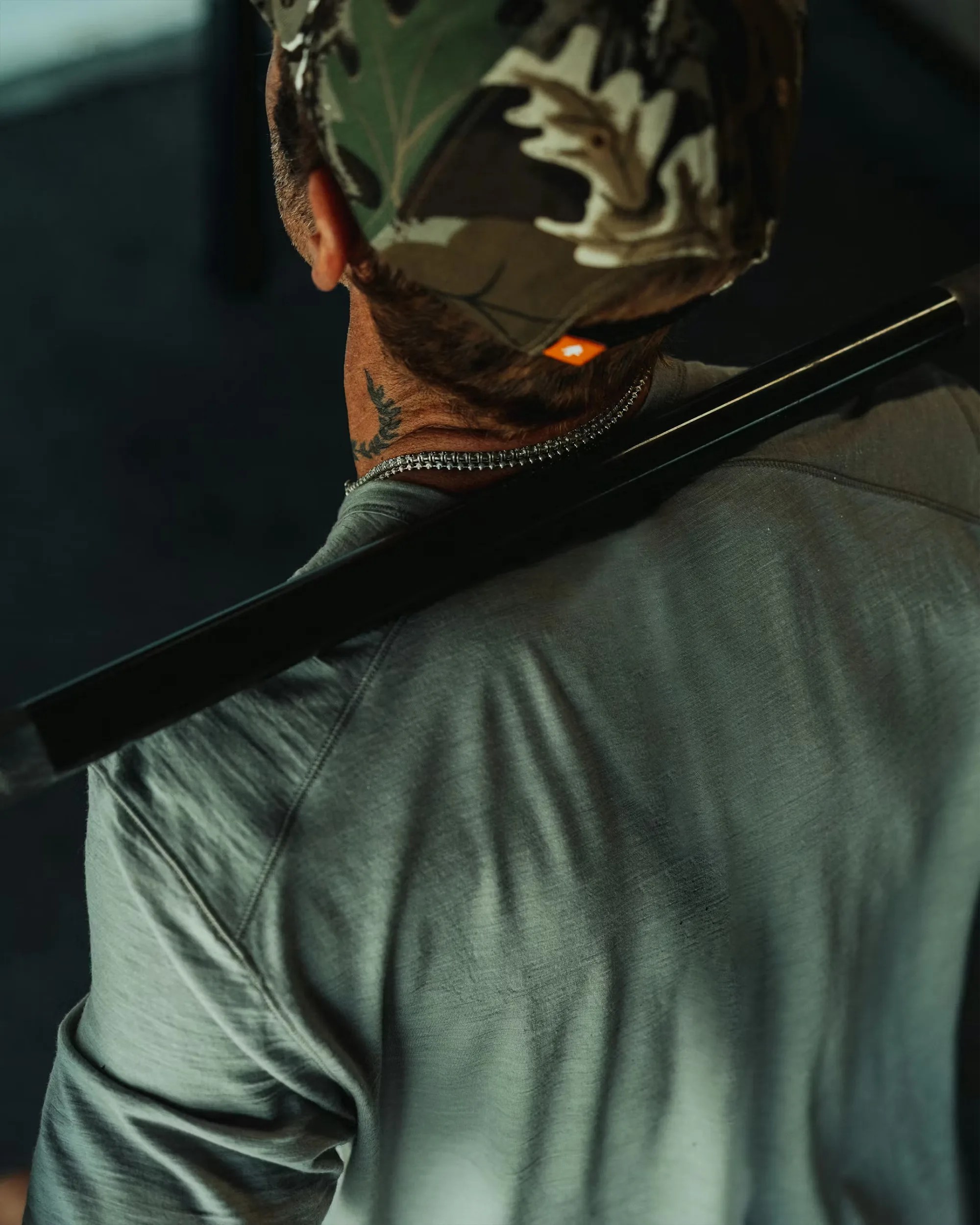 Man wearing a camouflage cap and gray shirt with a black bar resting on his shoulders.