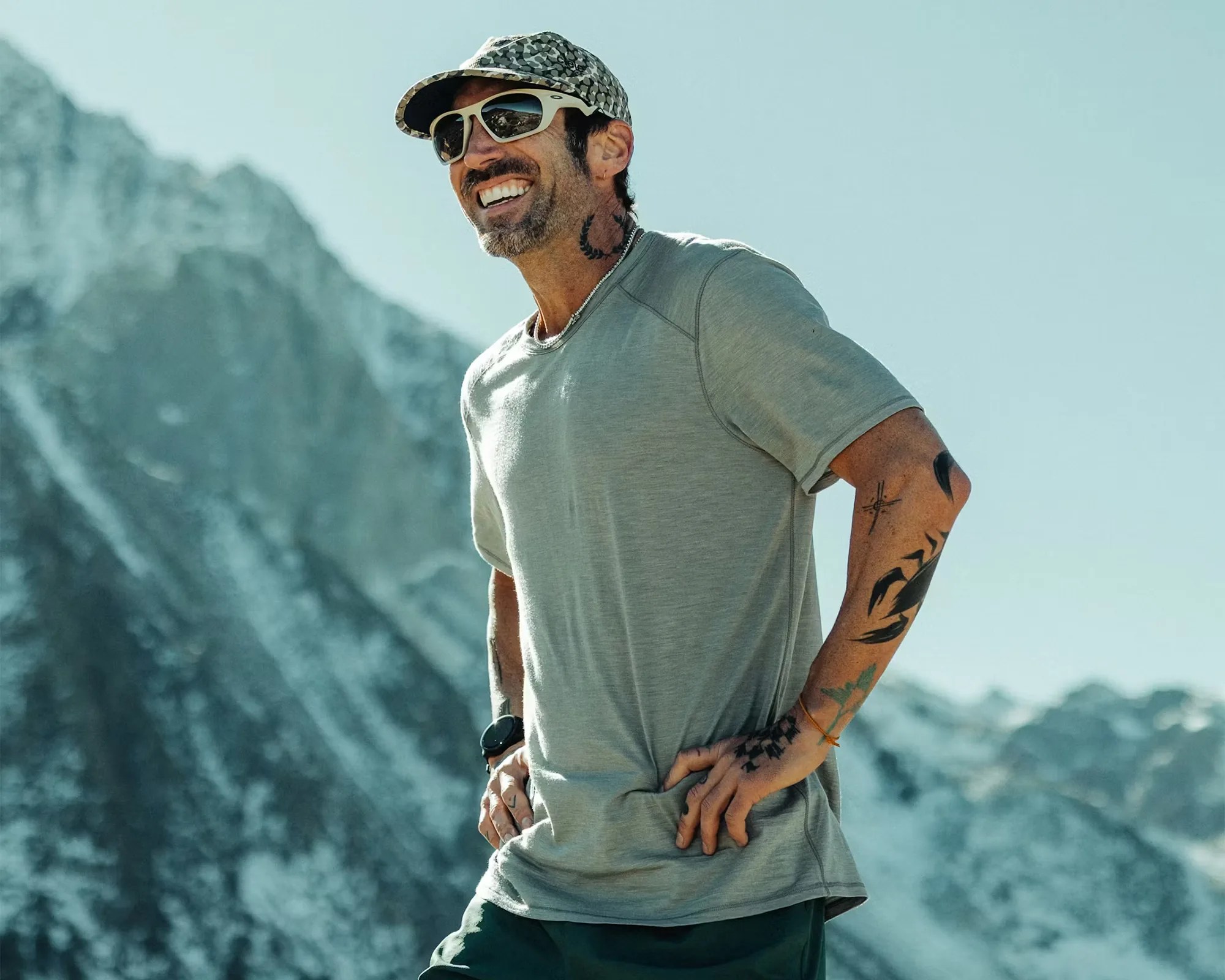 Man wearing a light gray short-sleeve shirt, patterned cap, white sunglasses, and a black wristwatch with mountain background.