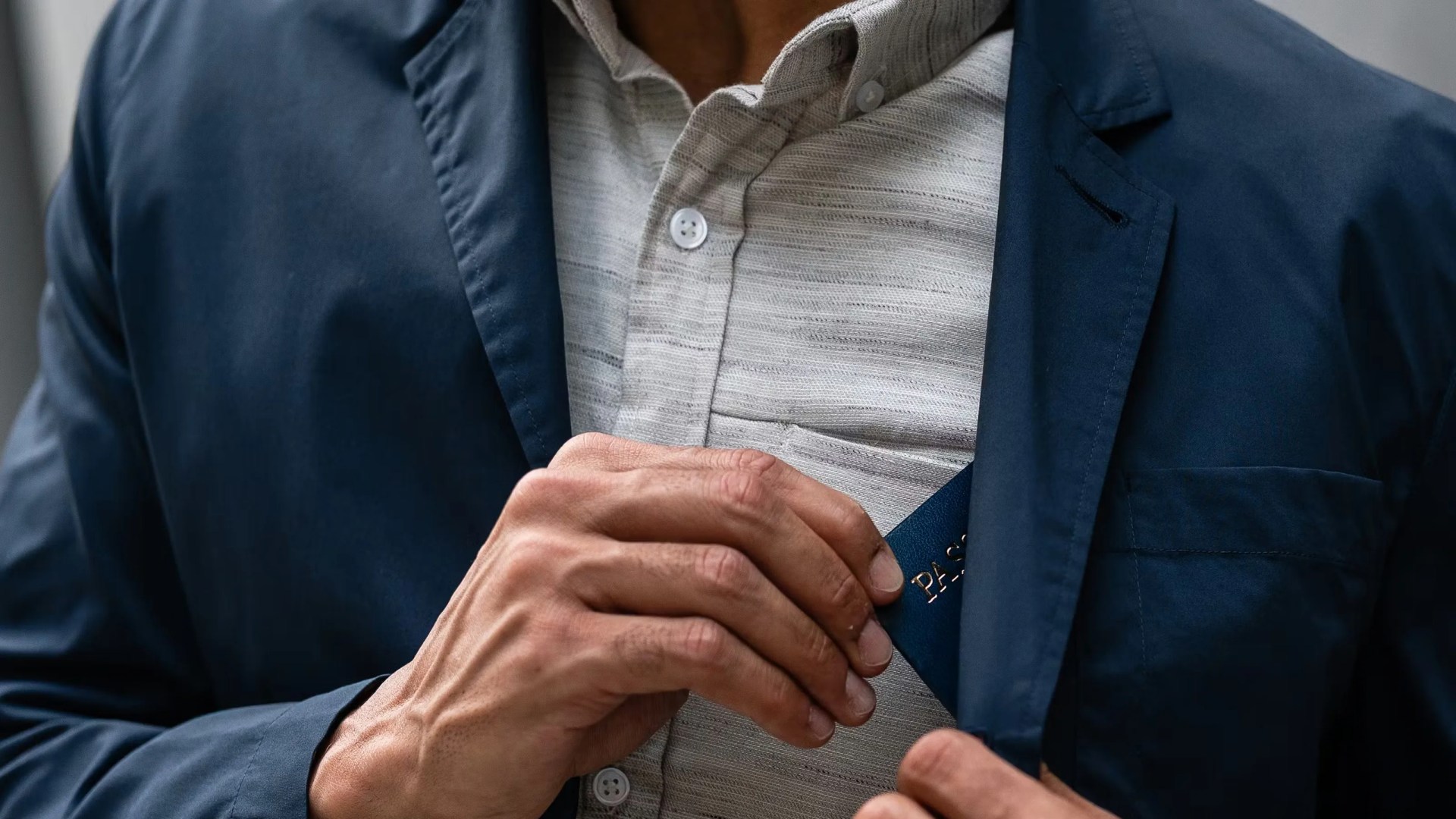 Hand placing a navy blue passport into the inner pocket of a navy blazer worn over a light gray shirt.