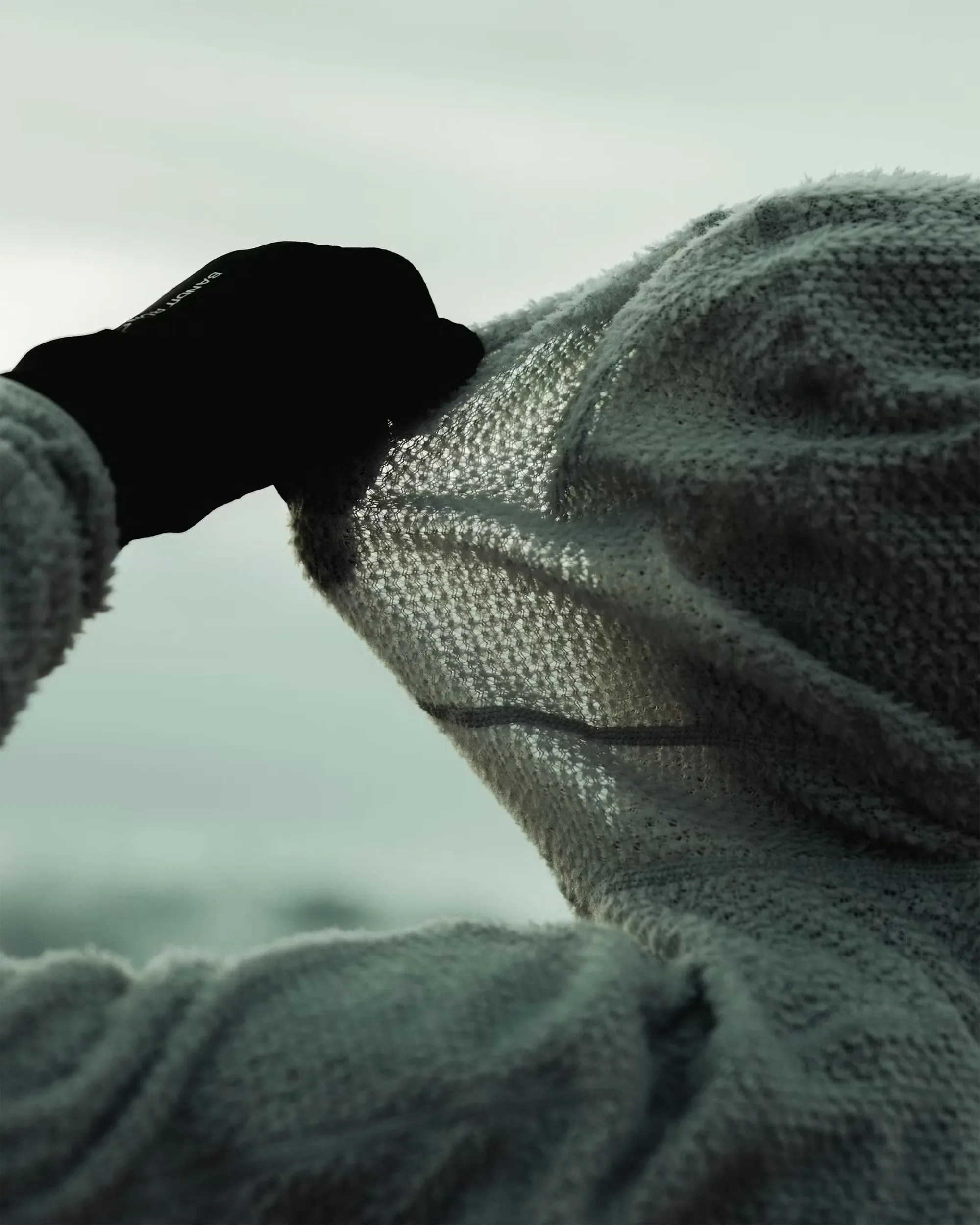 Person wearing a textured knit sweater and black glove holding up the sweater's hood against a cloudy sky.