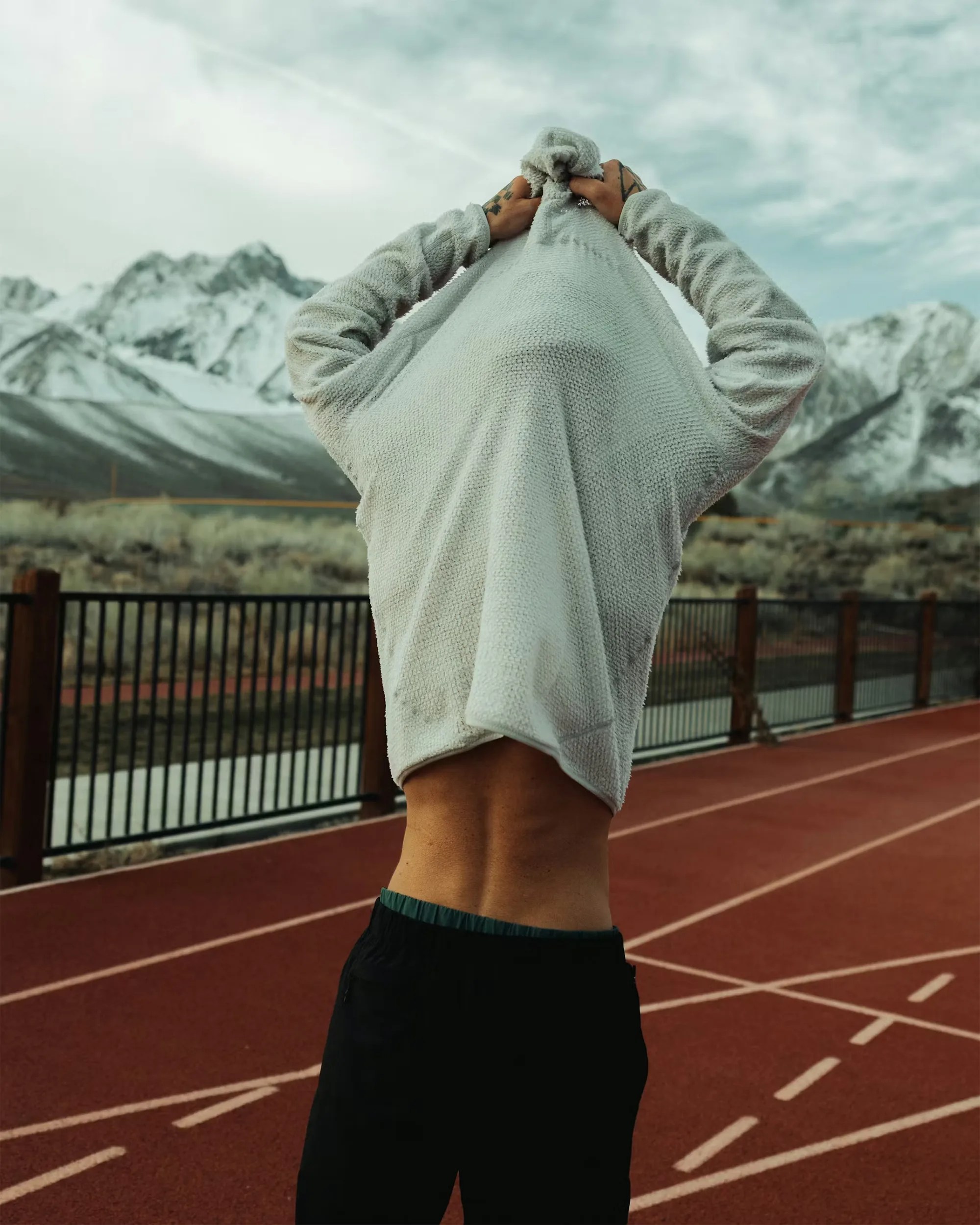 Person pulling a light gray sweater over their head on a red running track with snow-covered mountains in the background.
