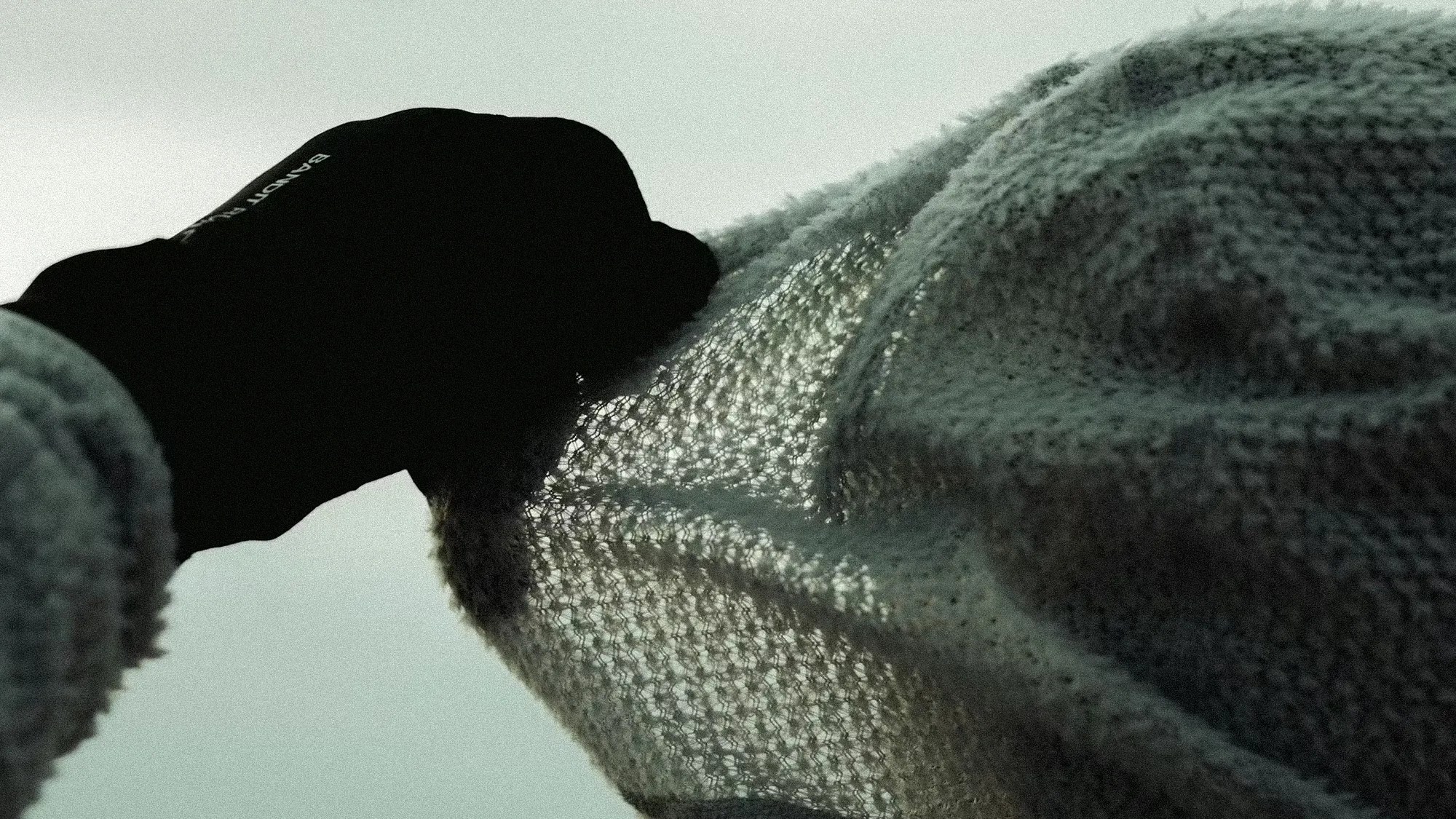 Person wearing a black glove holding up a loosely knit, light-colored fabric against a pale sky.