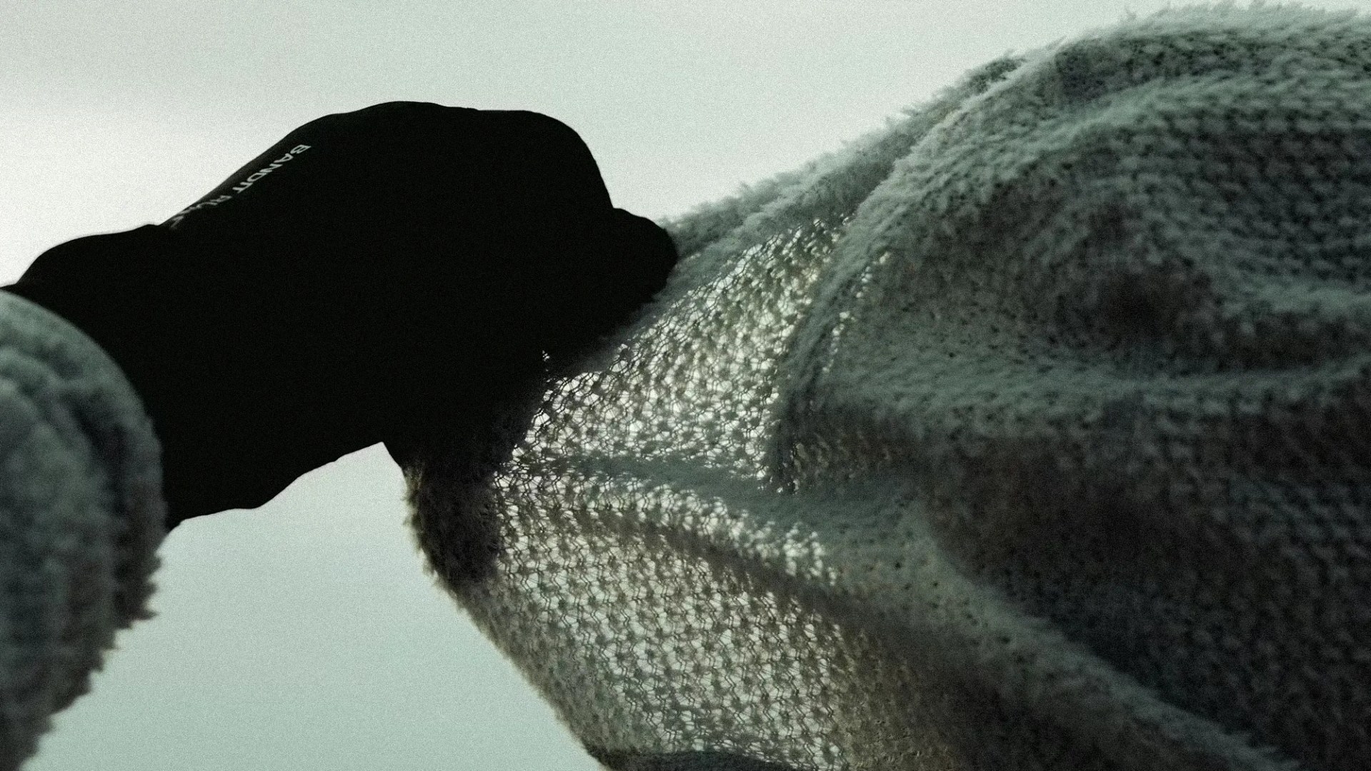 Person wearing a black glove holding up a loosely knit, light-colored fabric against a pale sky.