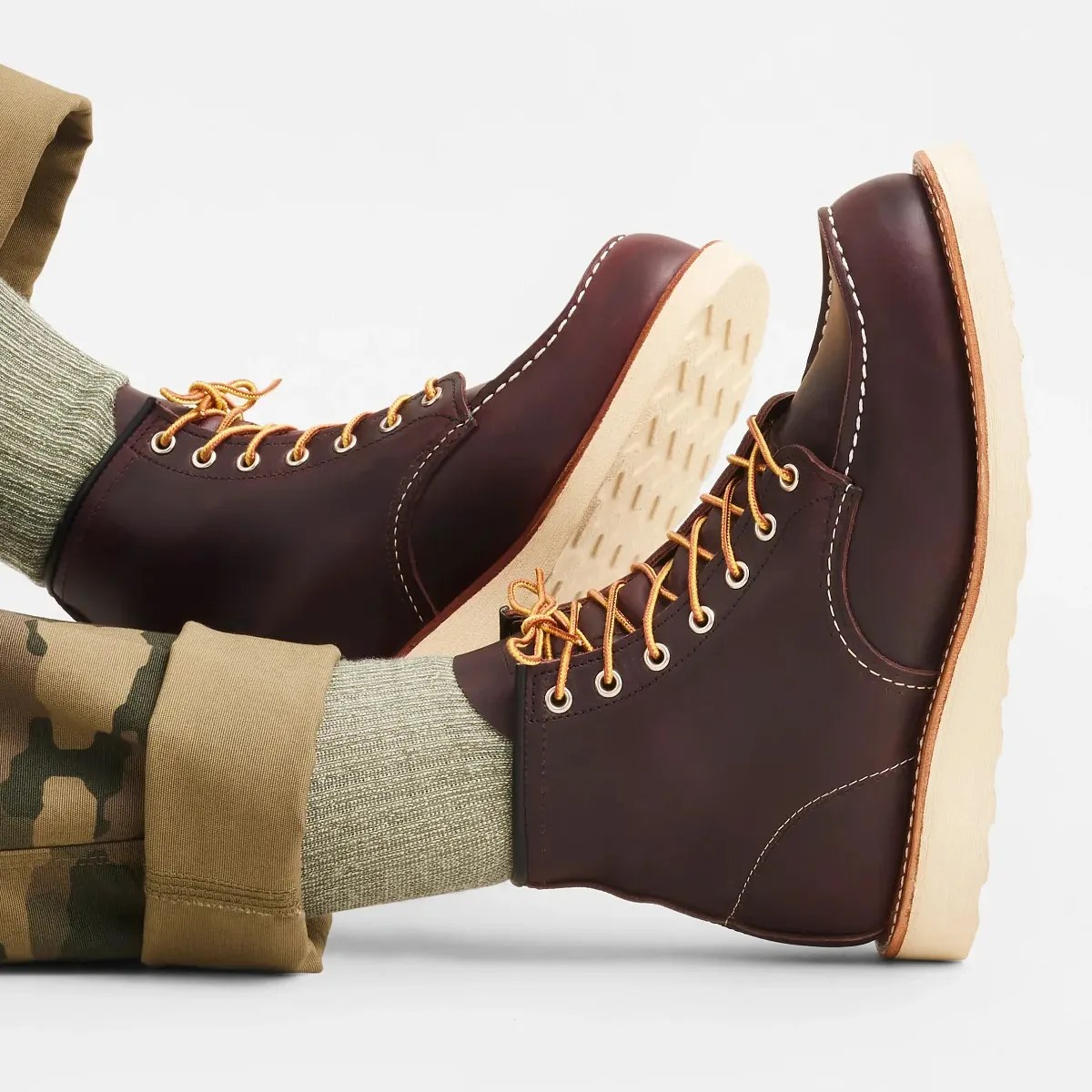 Pair of dark brown leather boots with yellow laces and light beige soles worn with green socks and camo pants.