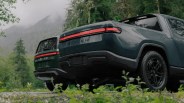 Two Rivian electric trucks, one dark green and one dark blue, parked on a forest road with misty mountains in the background.