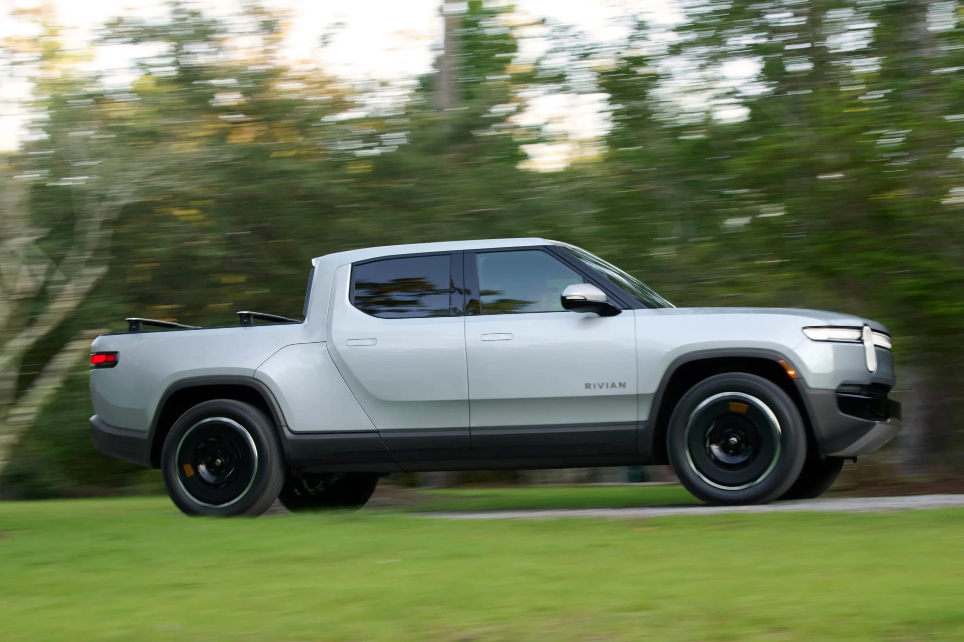 Silver Rivian electric pickup truck driving on a grassy road with blurred green trees in the background.