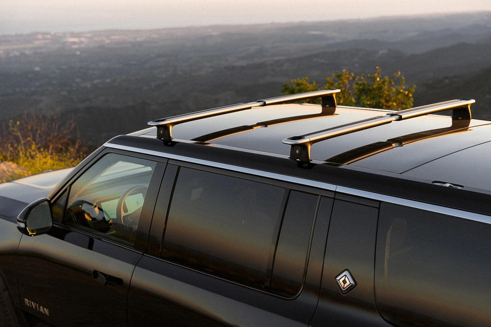 Black Rivian SUV with silver roof racks parked outdoors at sunset.