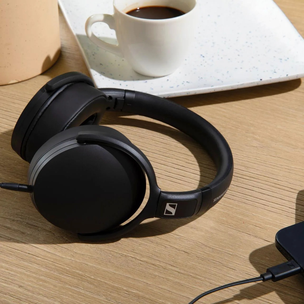 Black over-ear Sennheiser headphones with a cable connected, placed on a wooden table near a white coffee cup.