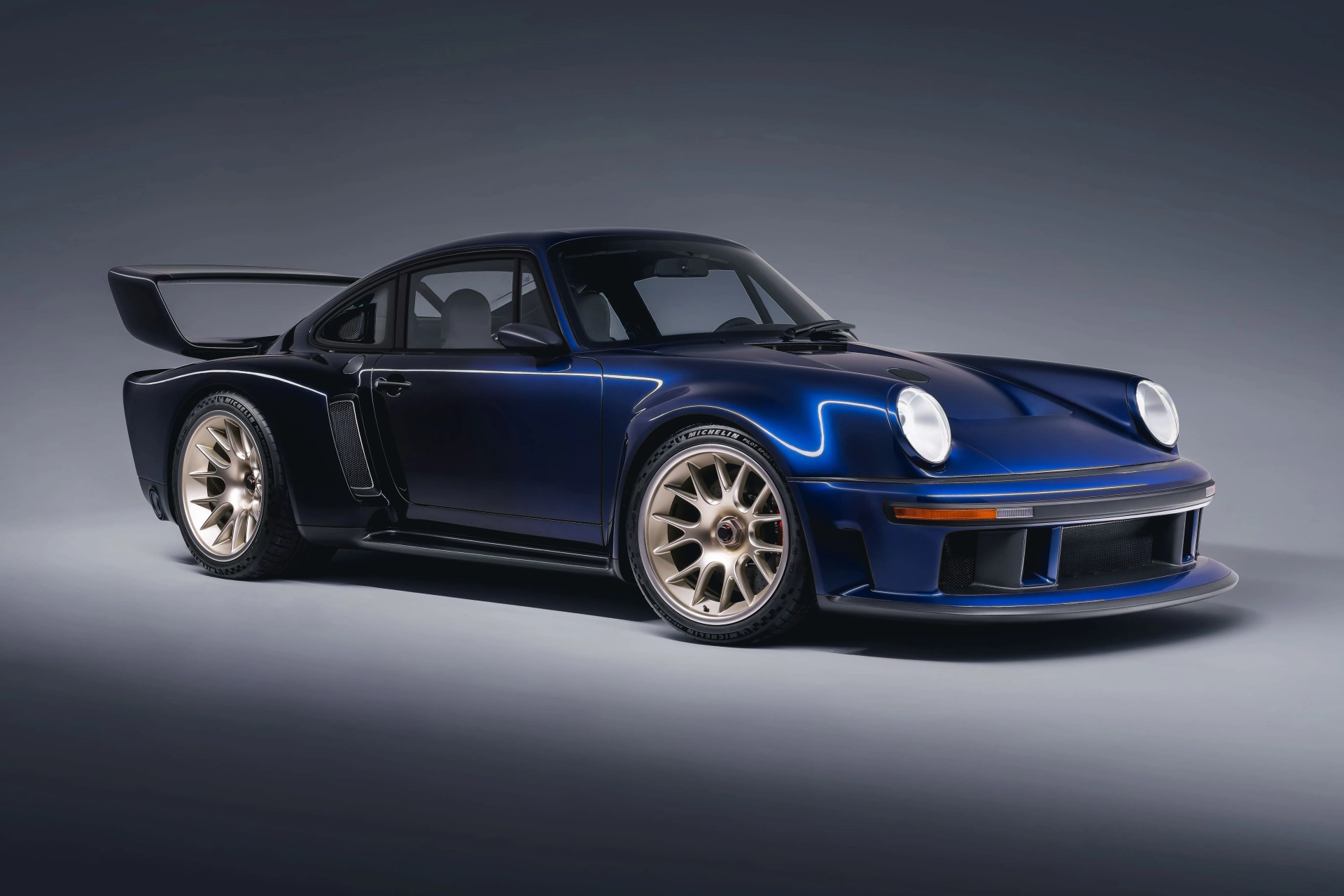 Dark blue classic sports car with wide body, large rear wing, and gold multi-spoke wheels in a studio setting.