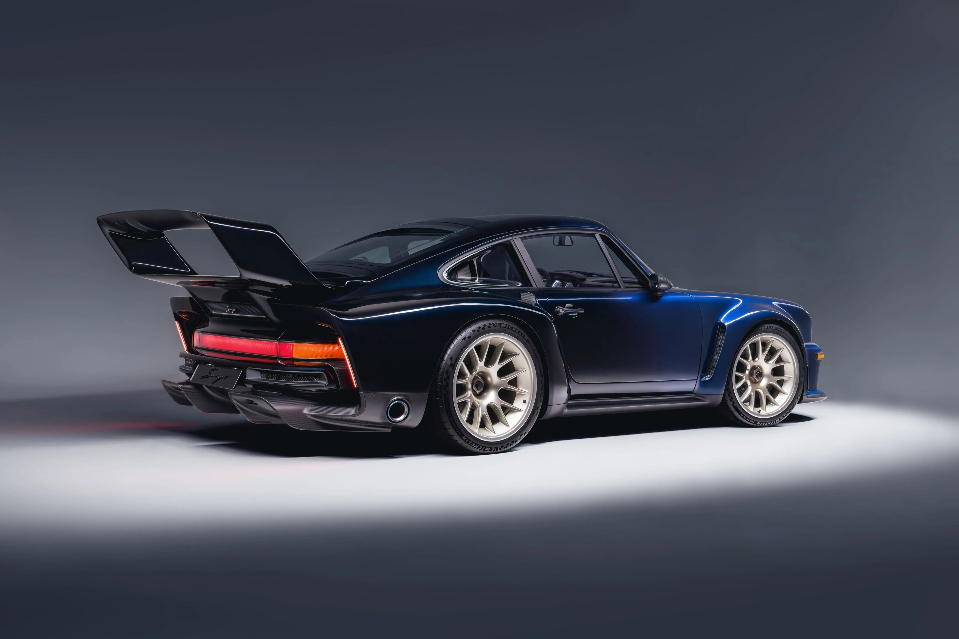 Dark blue classic sports car with large rear spoiler and gold multi-spoke wheels on a gray studio floor.