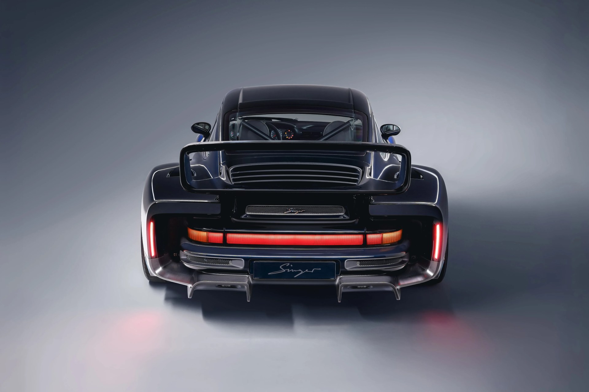 Rear view of a black Singer sports car with illuminated taillights and a large rear wing on a gray background.