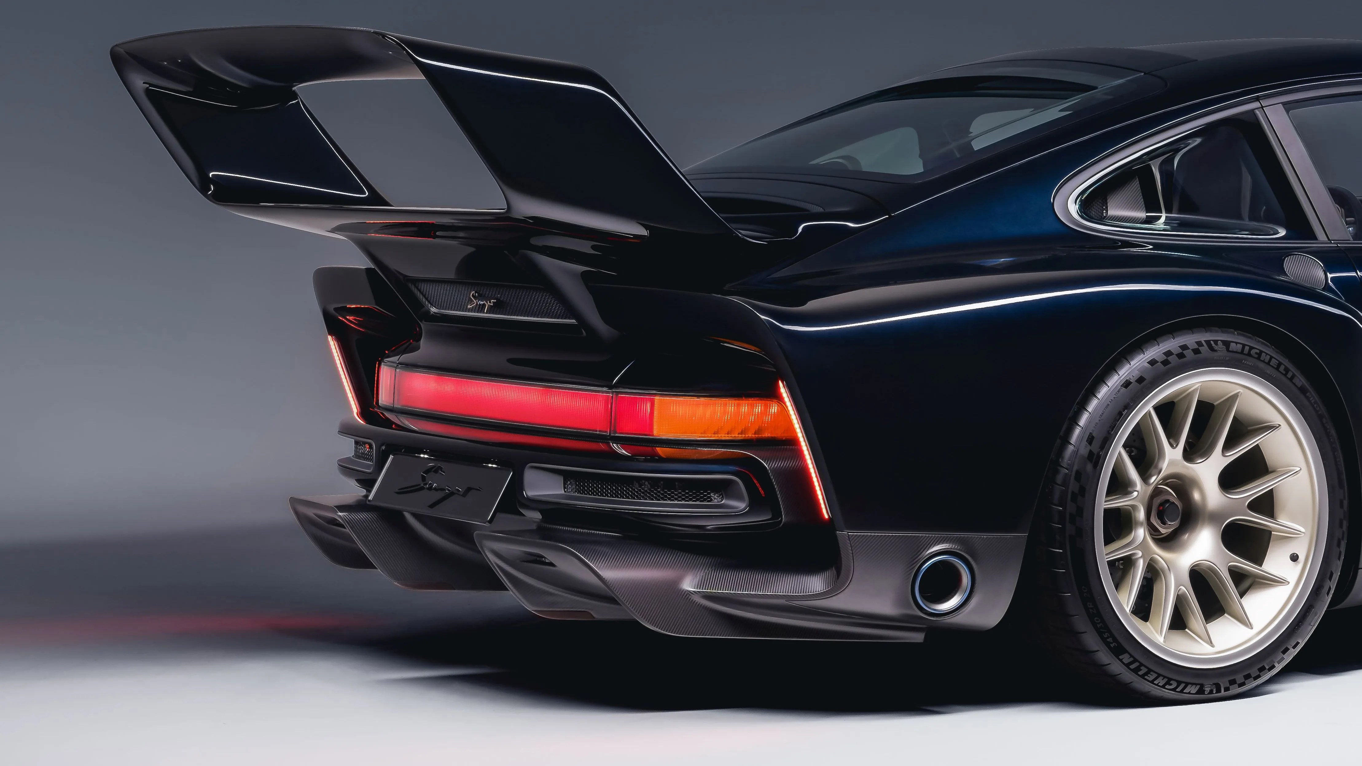 Rear view of a black Porsche 911 with large rear spoiler, red and orange taillights, and gold alloy wheel.