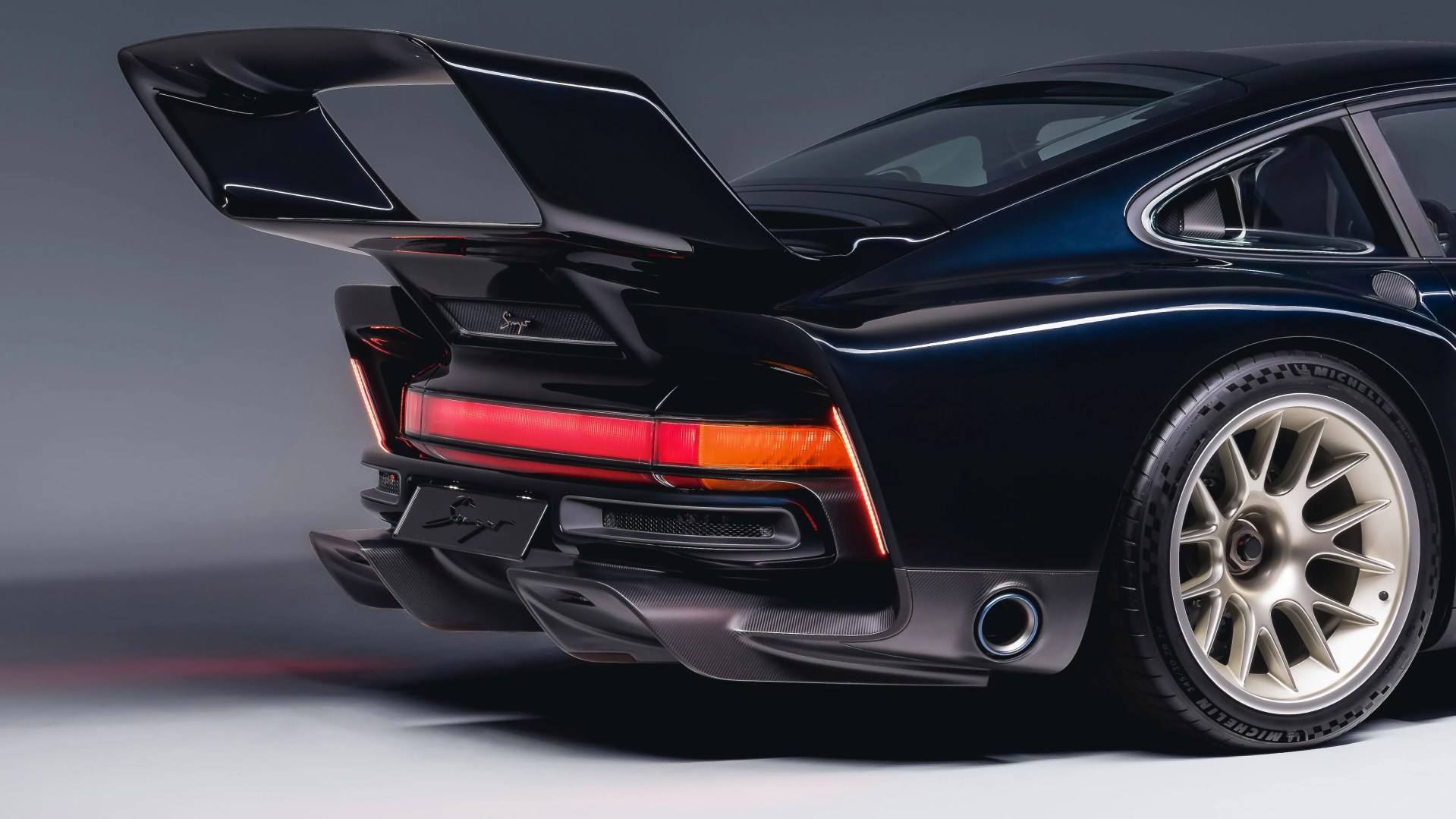Rear view of a black Porsche 911 with large rear spoiler, red and orange taillights, and gold alloy wheel.