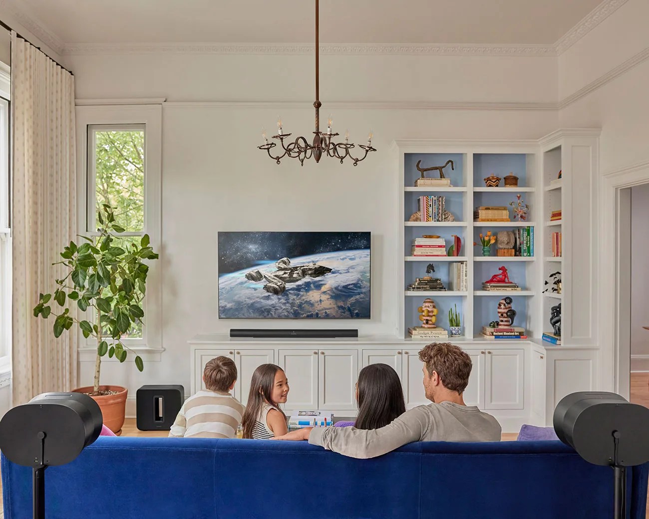 Family of four sitting on a blue couch watching a space-themed show on a wall-mounted TV in a bright living room.