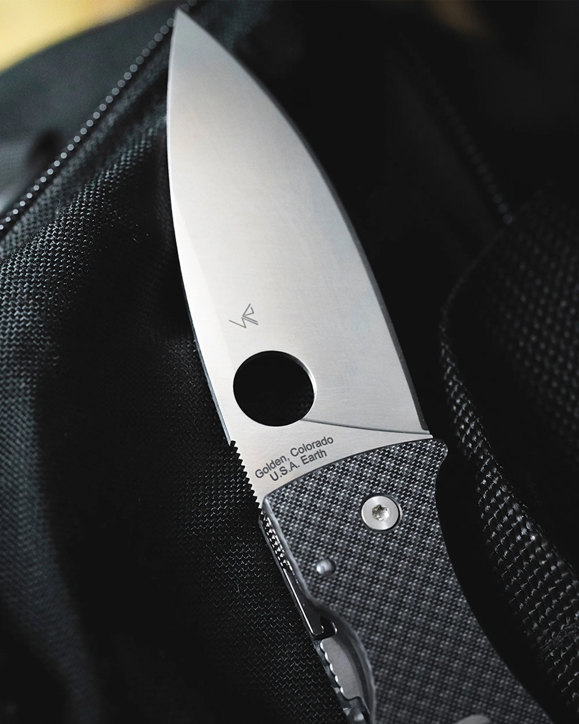 Close-up of a folding knife with a textured gray handle and a stainless steel blade engraved with "Golden, Colorado U.S.A. Earth.