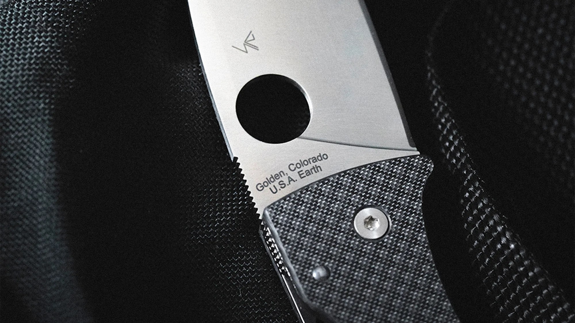 Close-up of a folding knife blade with a circular hole and textured handle, engraved with "Golden, Colorado U.S.A. Earth.