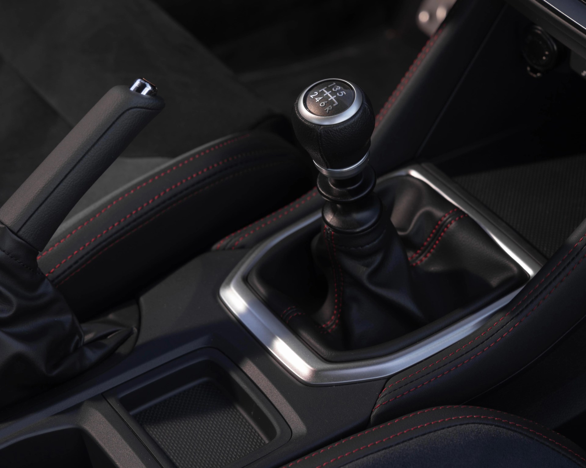 Black leather manual gear shift knob with silver trim and red stitching on car center console.