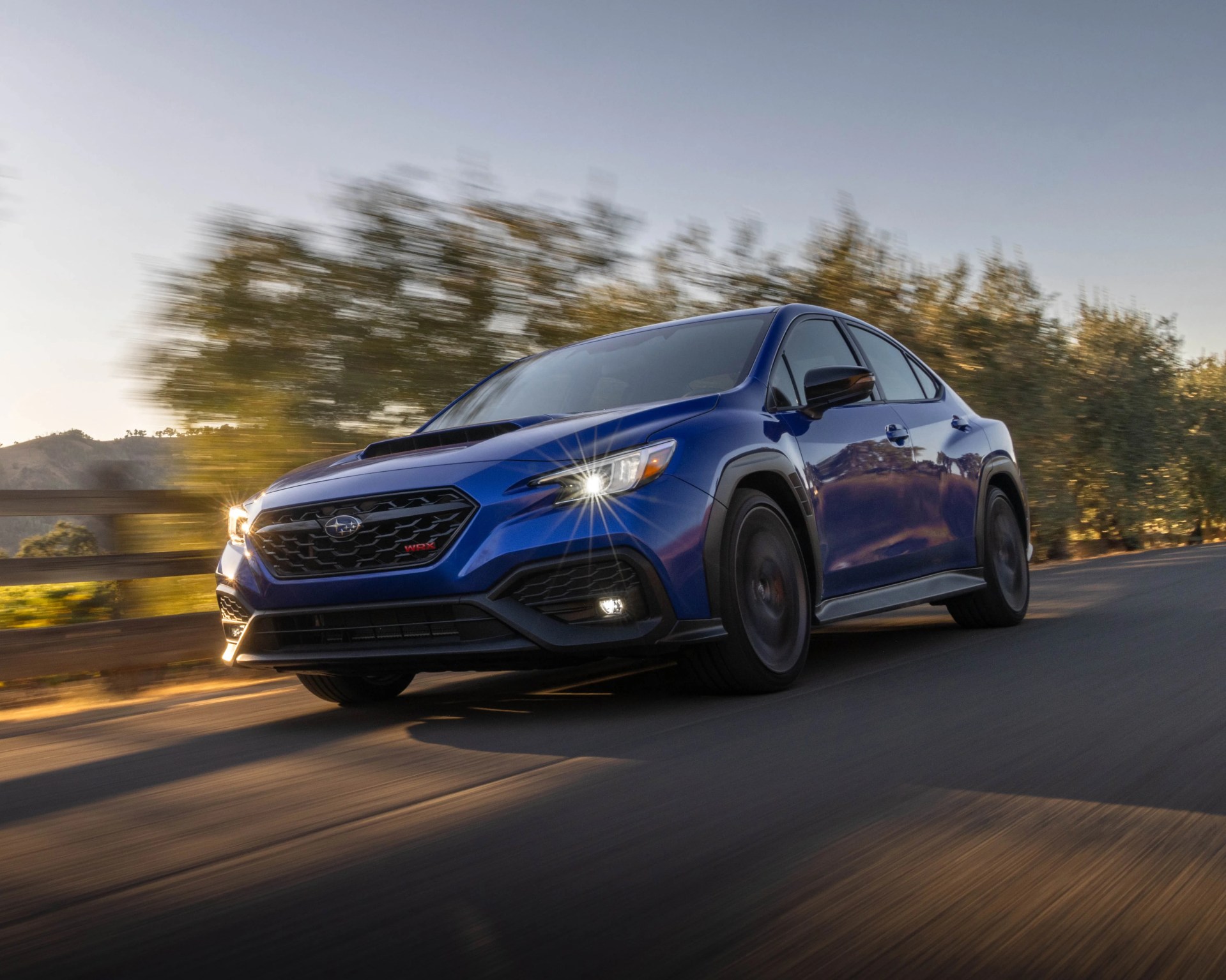 Blue Subaru WRX sedan with headlights on driving on a road with blurred trees in the background.