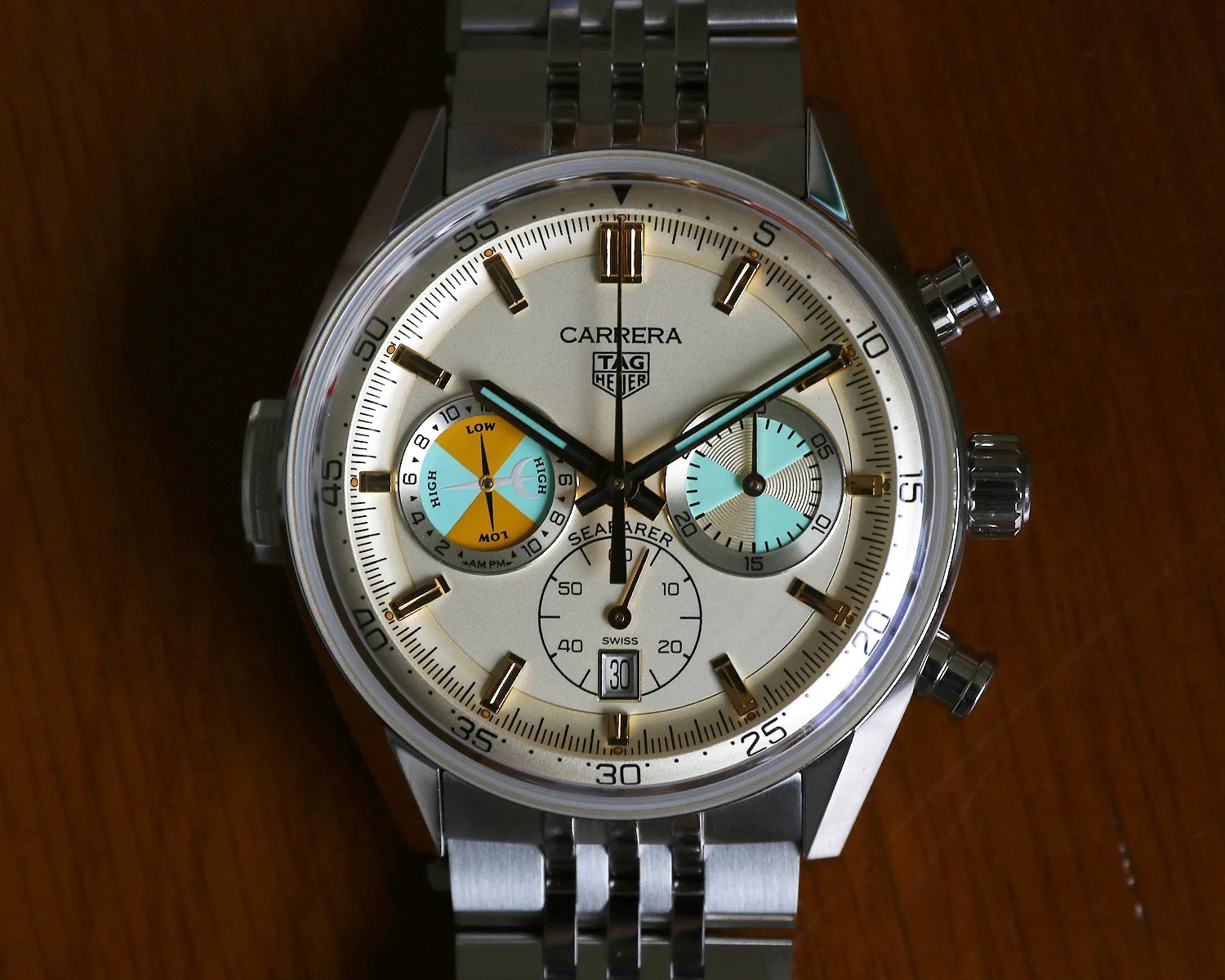 Silver TAG Heuer Carrera Seafarer wristwatch with white dial, gold hour markers, blue and orange subdials, and metal bracelet on a wooden surface.