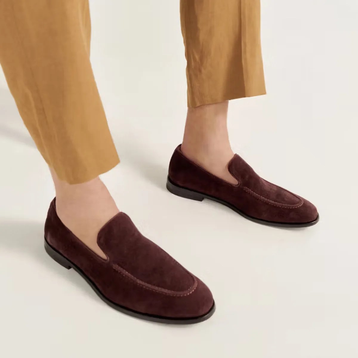 Pair of dark brown suede loafers worn with cropped tan pants on a light background.