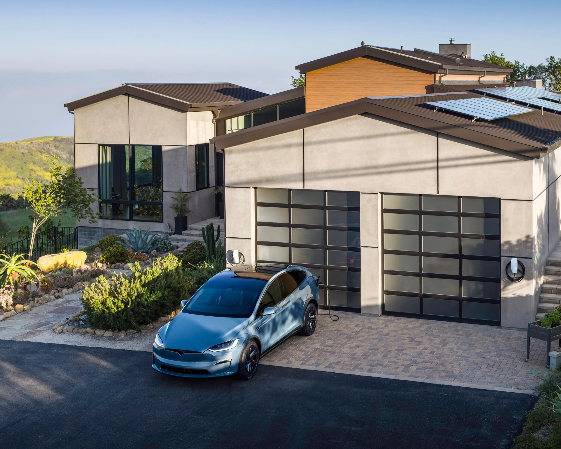 Blue Tesla Model X electric SUV charging in front of a modern house with solar panels on the roof.