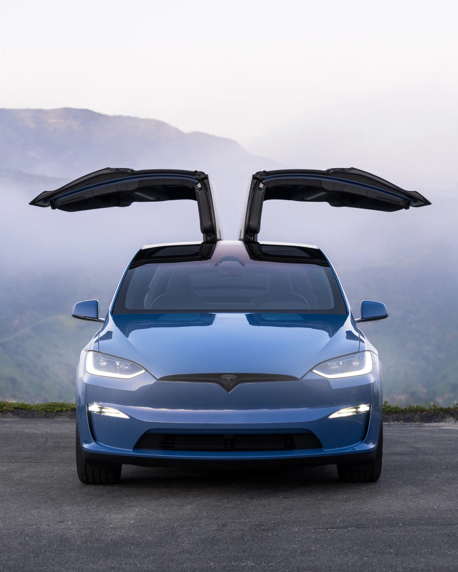 Blue Tesla Model X with falcon wing doors open, parked on a road with misty mountains in the background.