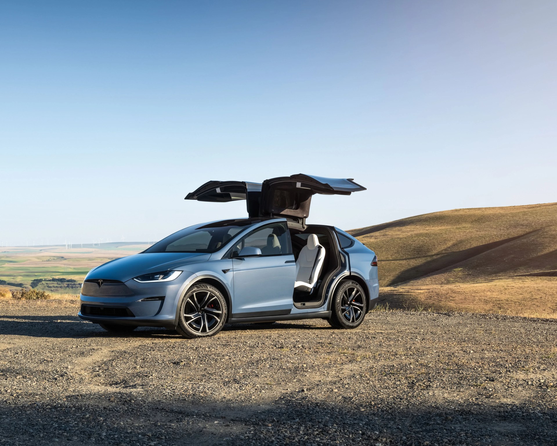Blue Tesla Model X with falcon-wing doors open on a gravel road with dry hills in the background.