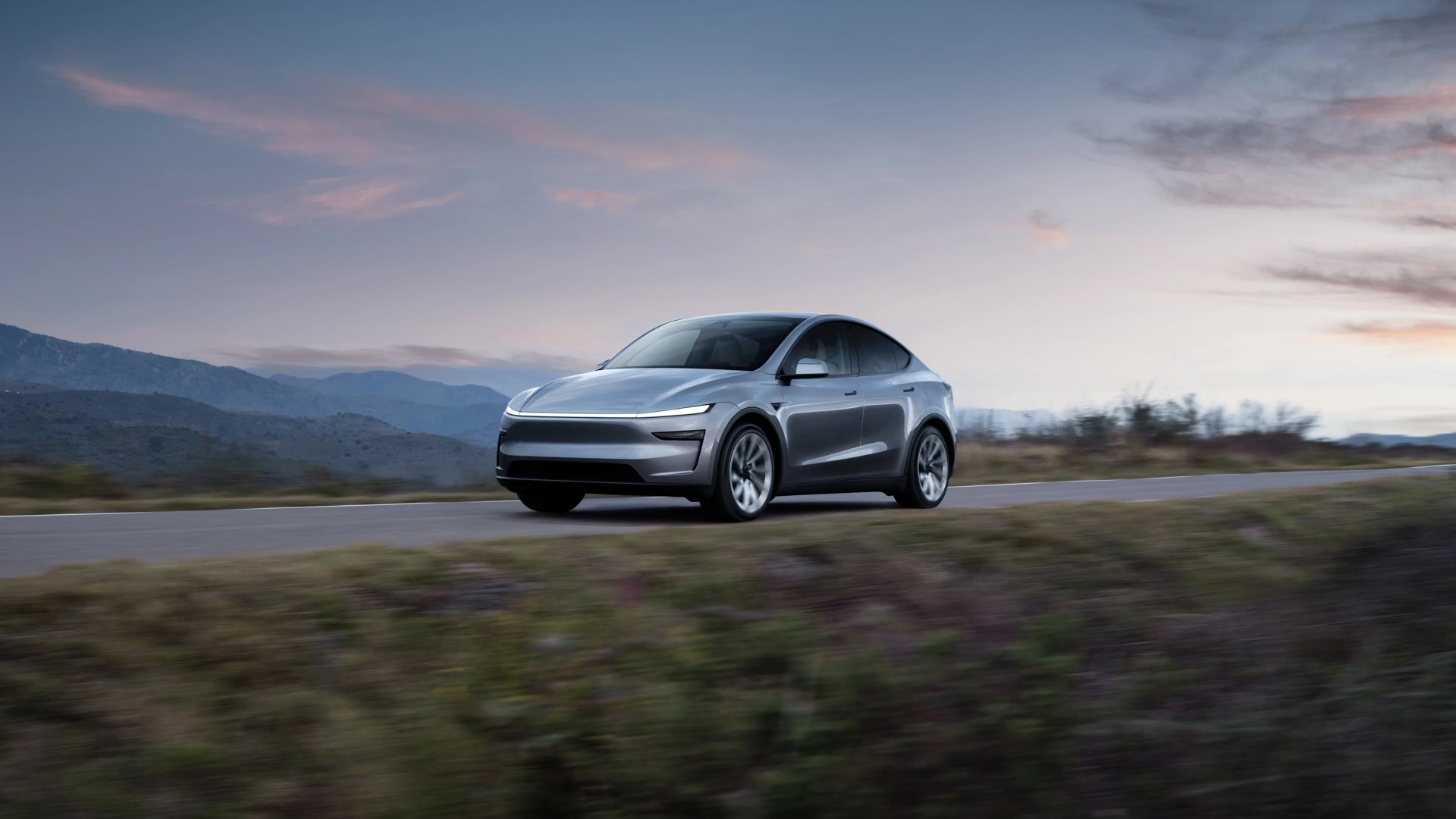 Silver electric SUV driving on a rural road with mountains and a cloudy sky in the background.