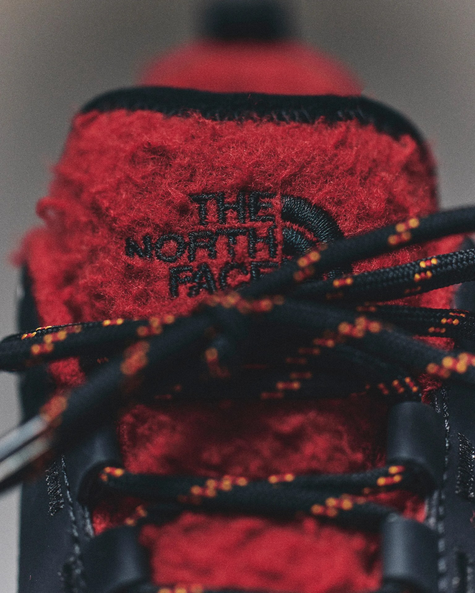 Close-up of red fuzzy fabric tongue on a black shoe with black and orange laces and The North Face logo embroidered in black.