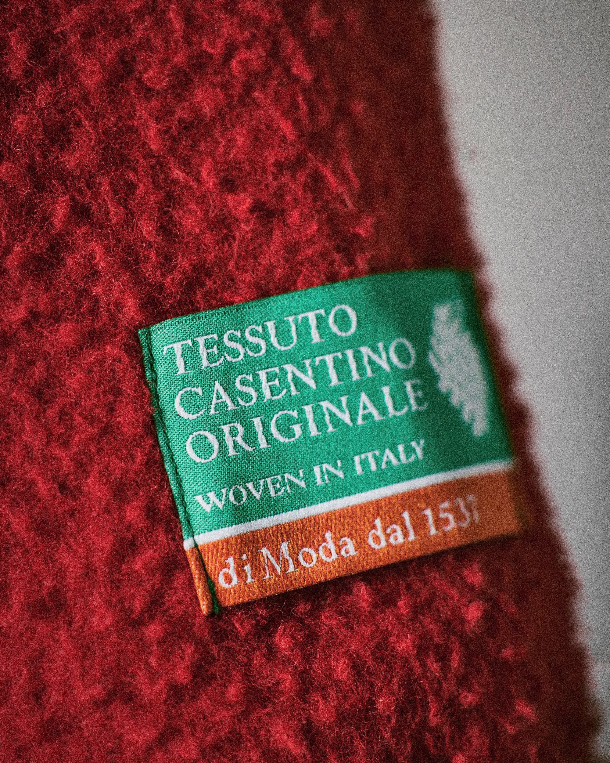 Green and orange fabric label with white text "Tessuto Casentino Originale Woven in Italy di Moda dal 1531" on red textured wool fabric.