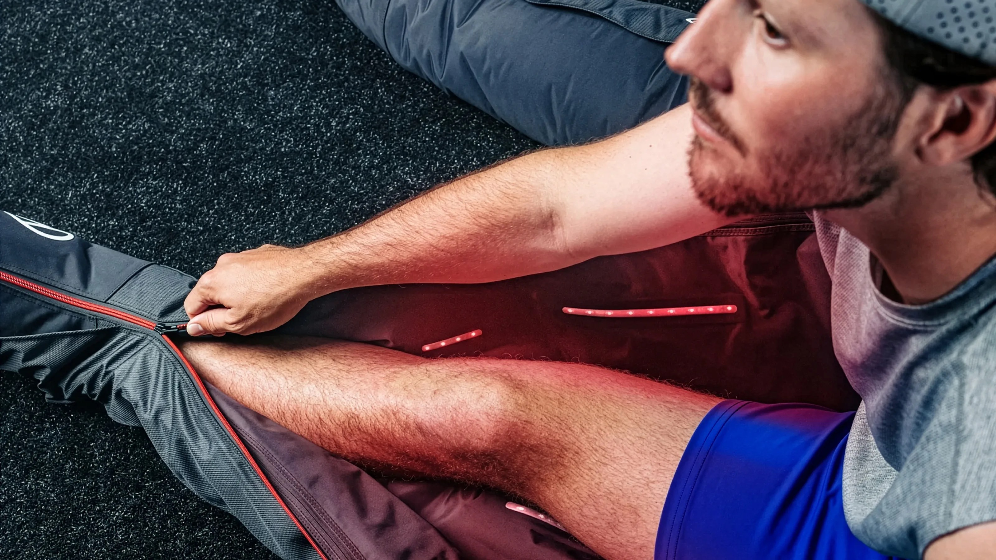 Man wearing blue shorts using a black and red compression recovery device on his leg while sitting on a carpeted floor.