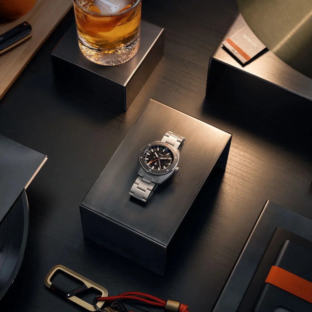 Silver metal wristwatch with black dial and orange second hand on a black display stand.