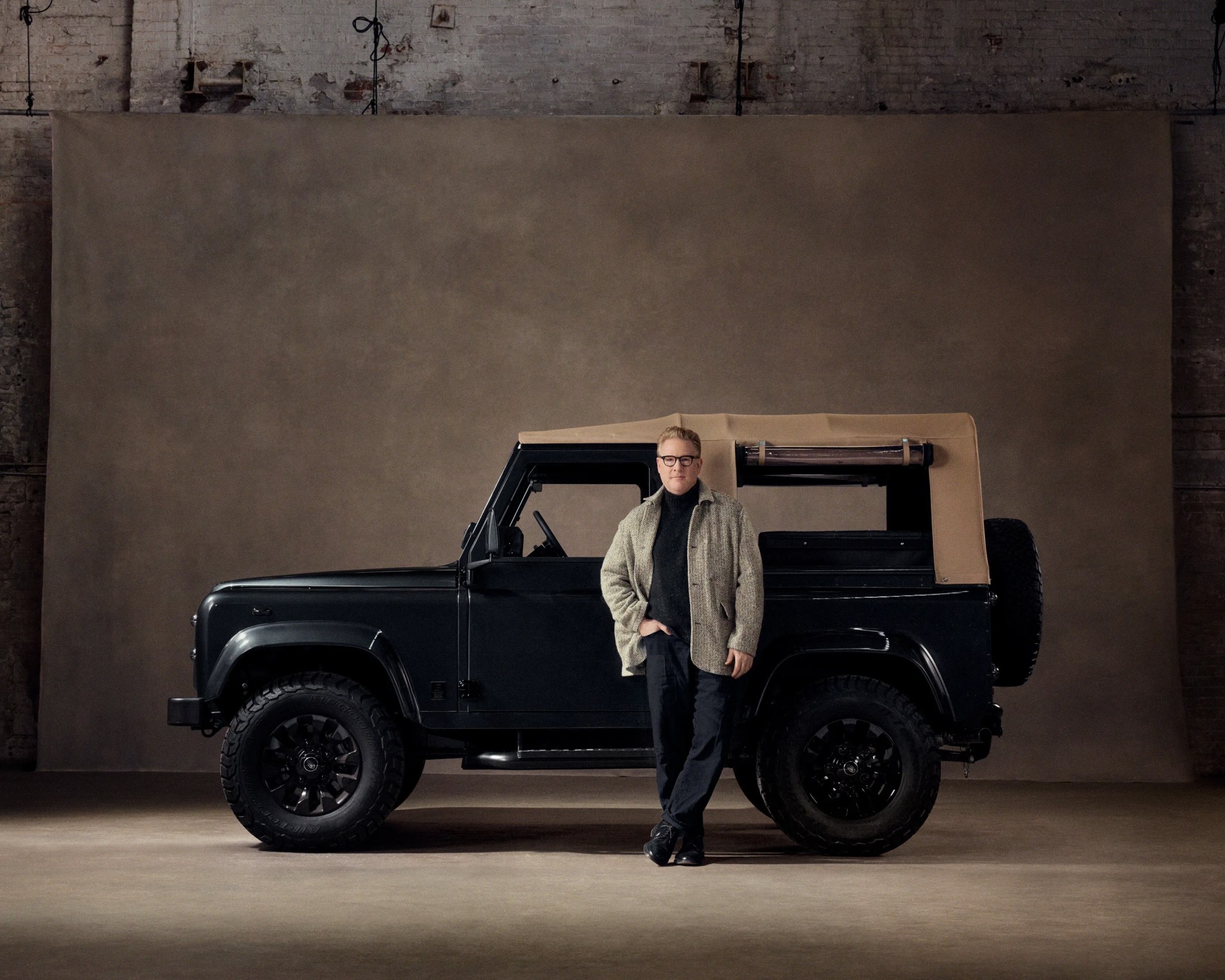 Man wearing glasses and a beige jacket standing in front of a black off-road vehicle with a tan soft top in an industrial setting.