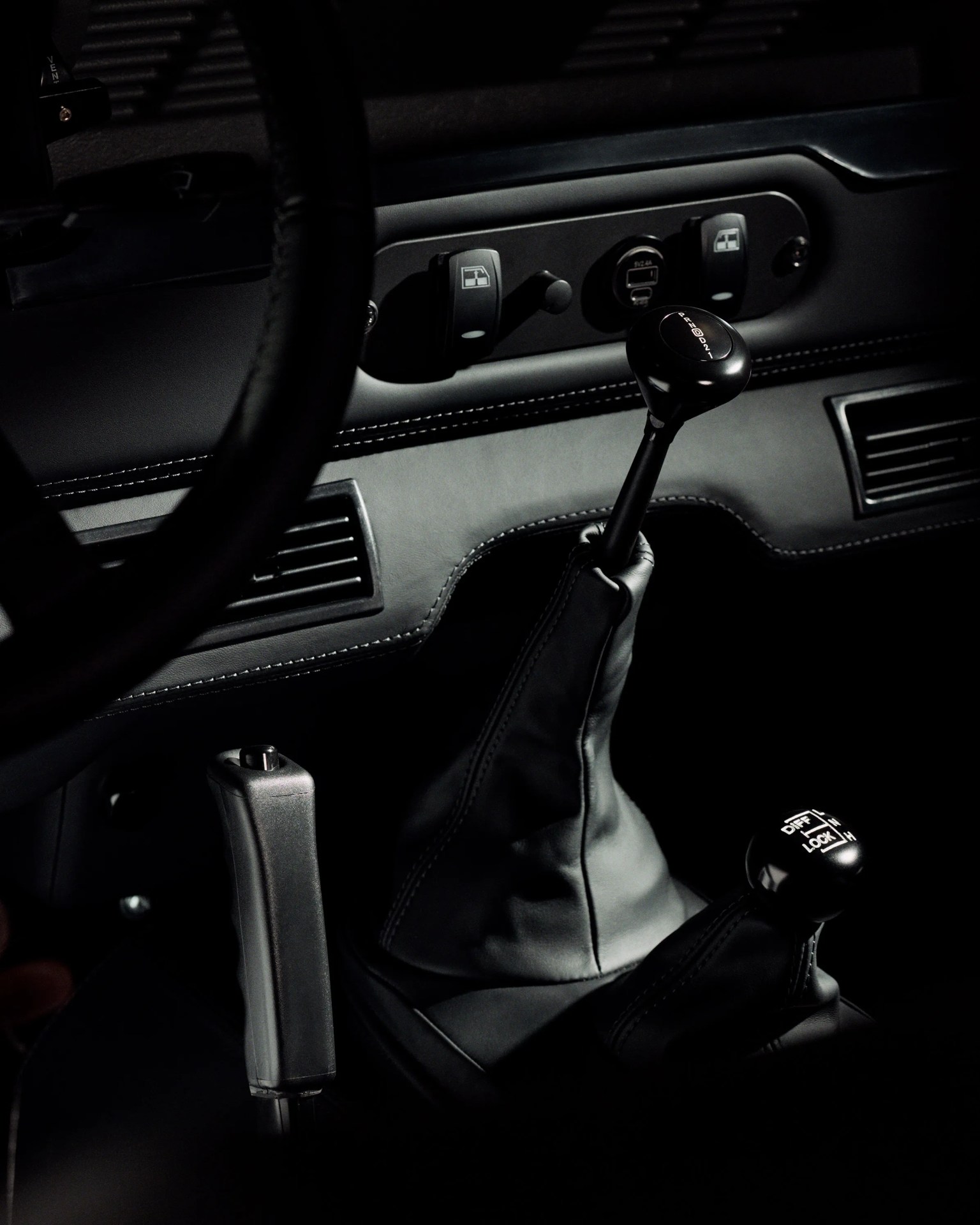 Black leather gear shift and handbrake in a dark car interior with visible dashboard controls.