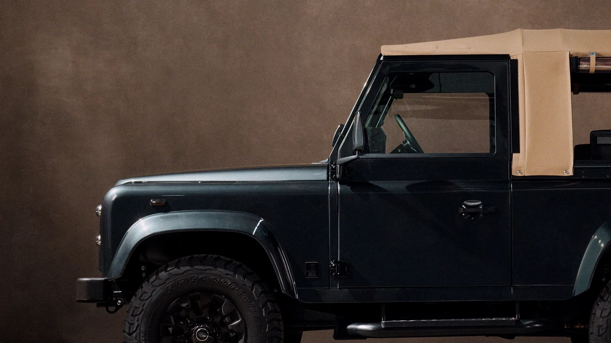 Dark green off-road vehicle with beige soft top and large black tires against a brown background.