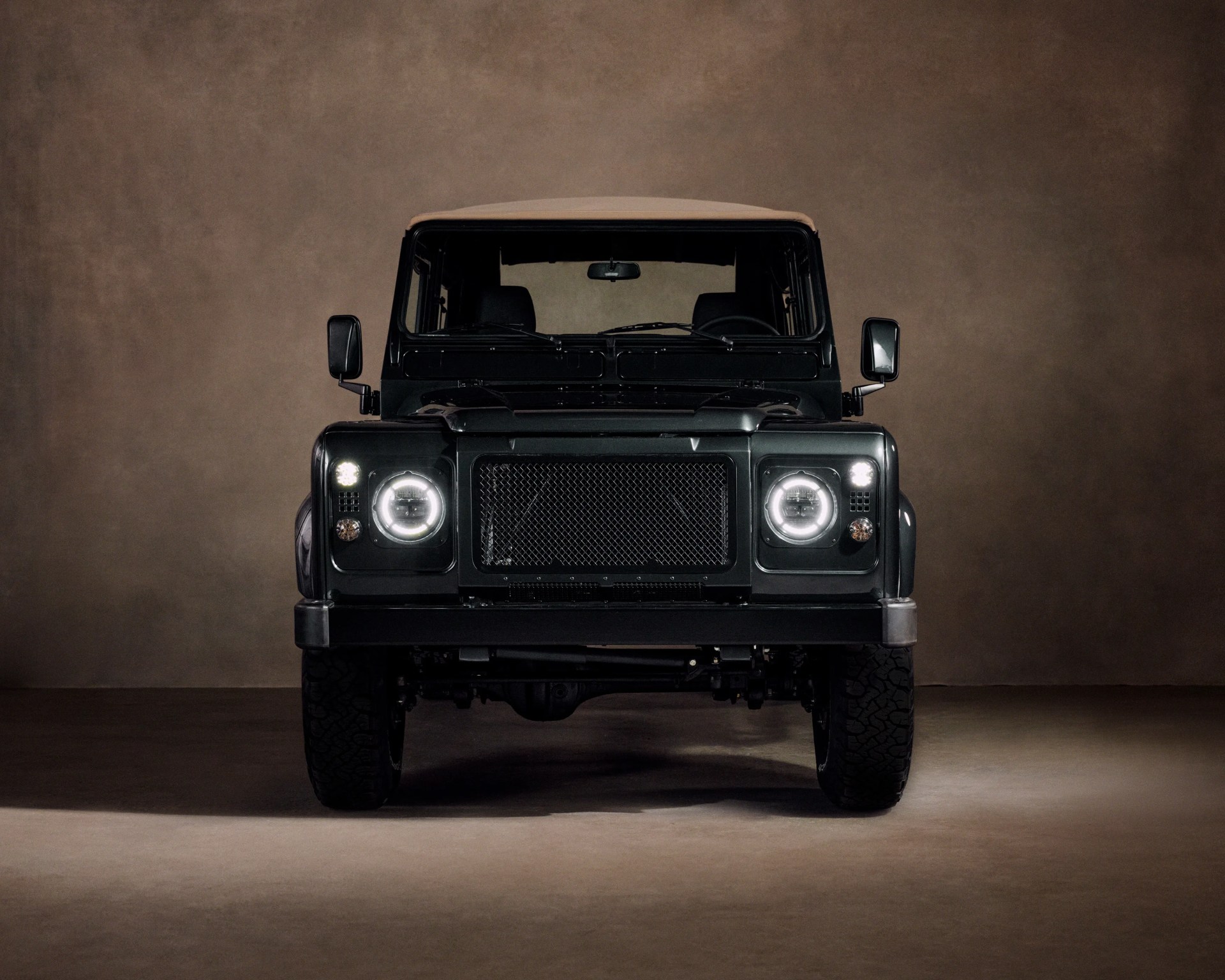 Front view of a dark green off-road vehicle with round LED headlights and a beige roof against a brown backdrop.