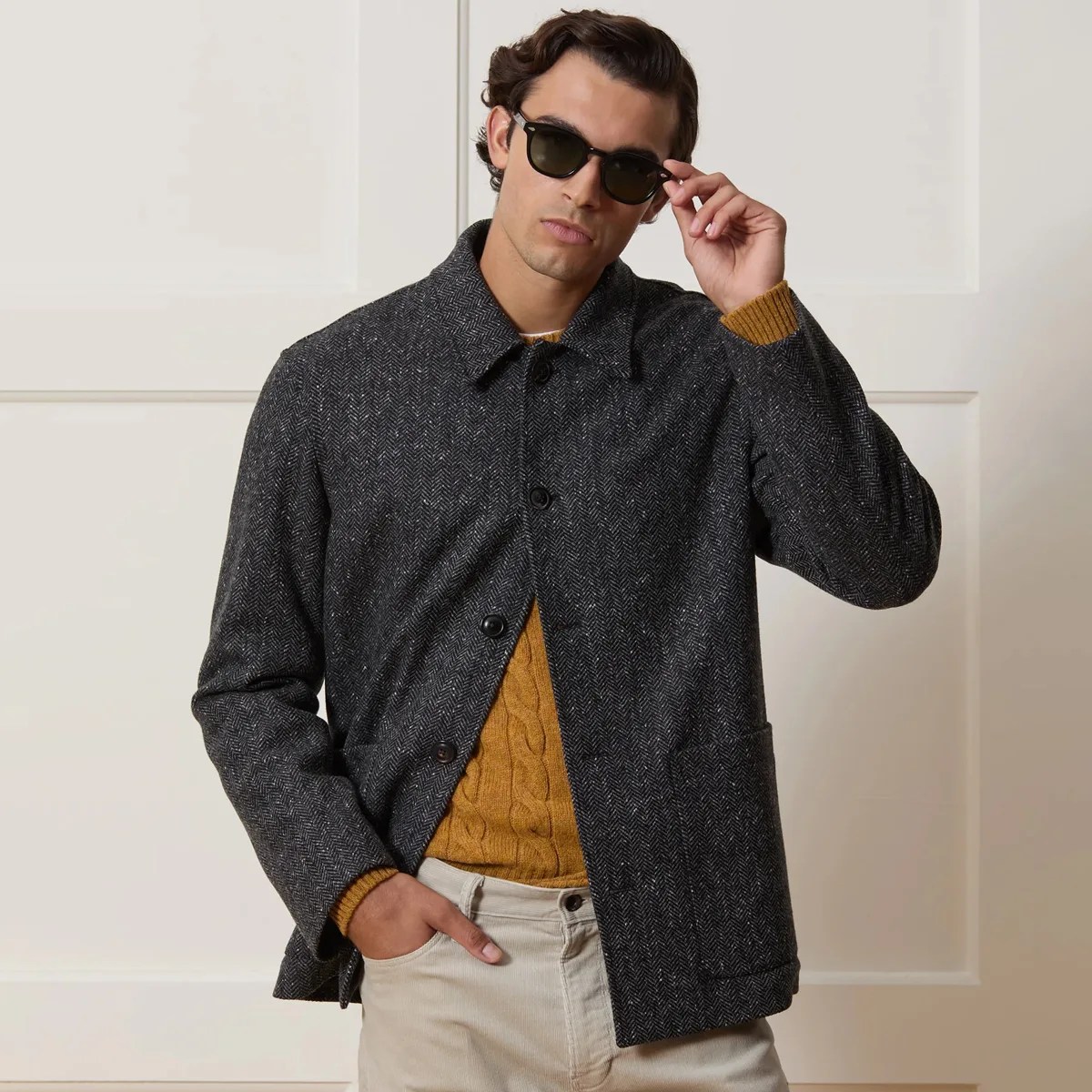 Man wearing a dark gray herringbone wool jacket over a mustard cable-knit sweater and beige pants, adjusting black sunglasses.