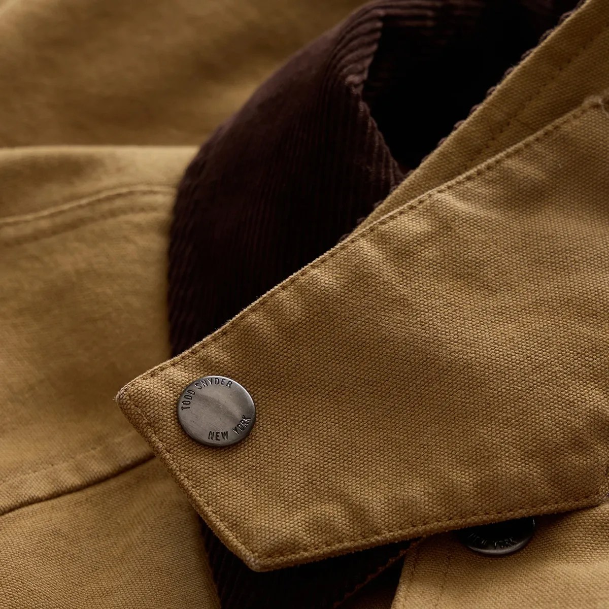 Close-up of a tan canvas jacket collar with a metal snap button engraved "Todd Snyder New York" and dark brown corduroy fabric underneath.