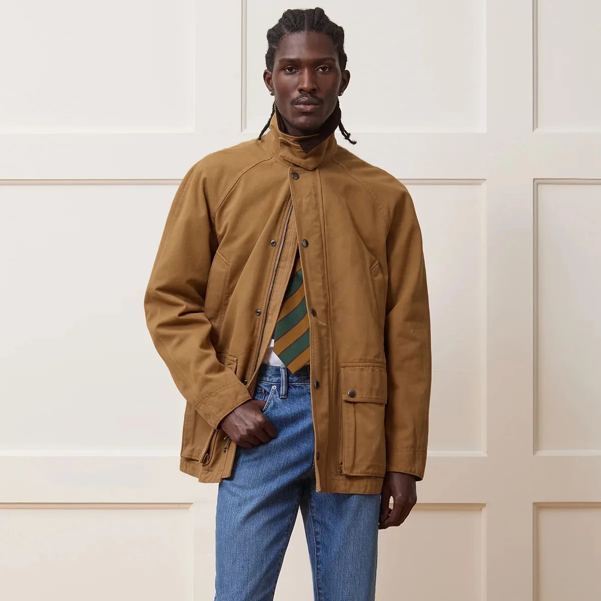 Man wearing a tan zip-up jacket with snap buttons and blue jeans against a paneled wall.