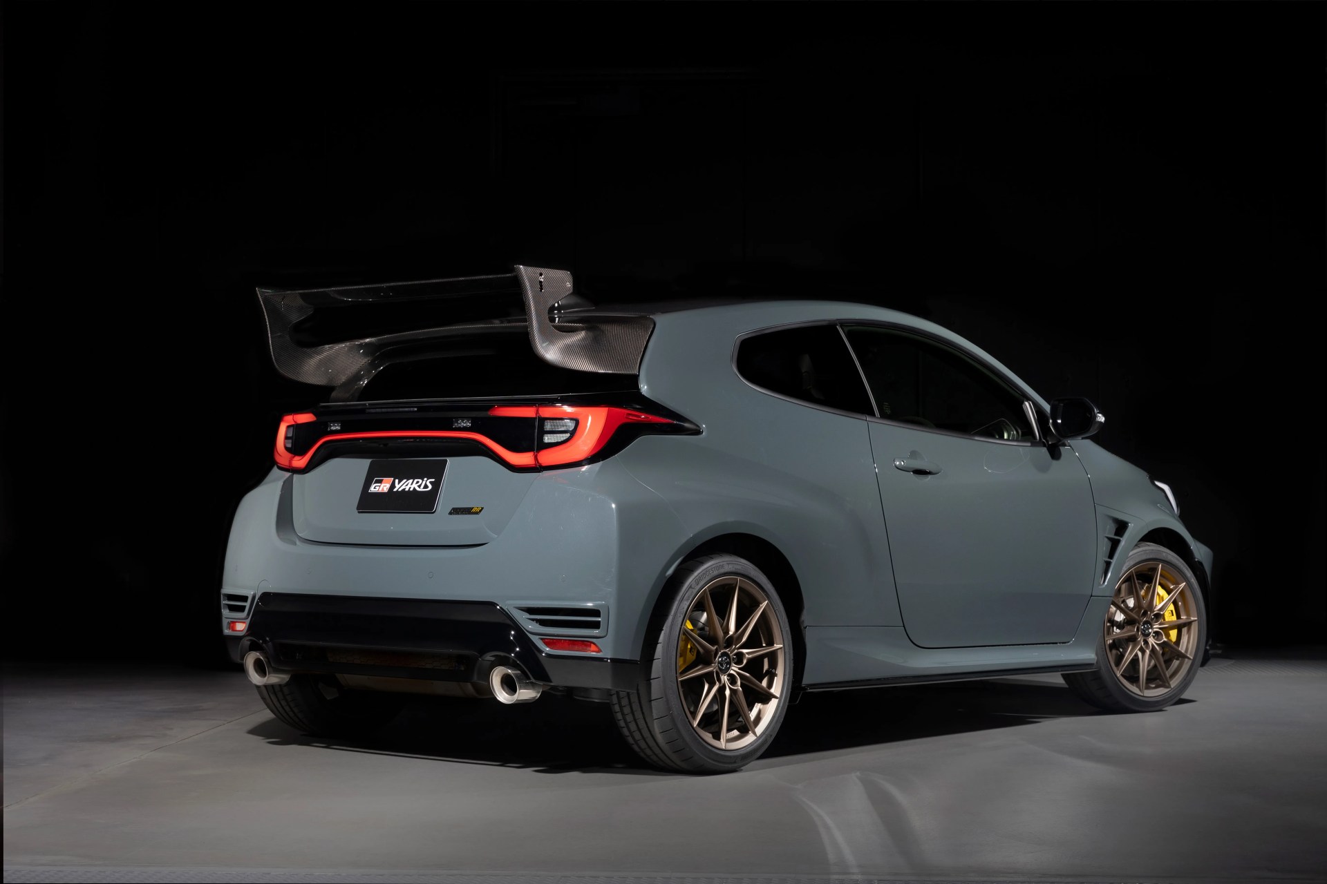 Gray Toyota GR Yaris hatchback with large carbon fiber rear wing and bronze alloy wheels in a dark studio.